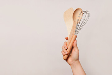 Hand holding a wooden spoon, a wooden spatula, and a whisk against a plain beige background, copy space