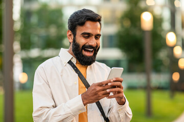 Indian bearded man is happily engaged with his smartphone while standing in a lush green park...