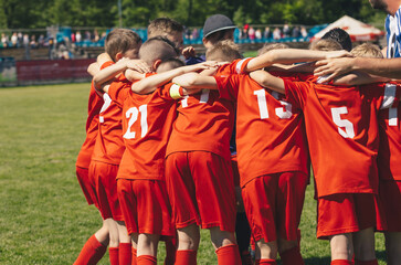 Children's sports team with coach. Kids play sports match on stadium field. School boys in red jersey shirts huddling in a team. Parents sitting at the stadium and watching pupils in the background 