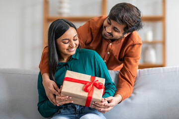 Indian man sitting on a couch is handing a gift to a woman next to him. The woman appears surprised...