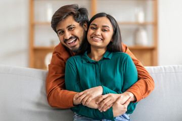 A smiling Indian couple is sitting on a comfortable sofa, warmly embracing each other in a well-lit...