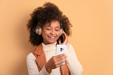 joyful black young woman listening to music with headphones in beige studio background. leisure, fun, music, dance concept.