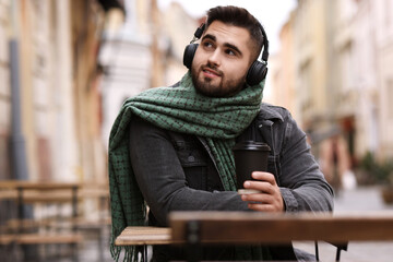 Fototapeta premium Smiling man in warm scarf with paper cup listening to music in outdoor cafe