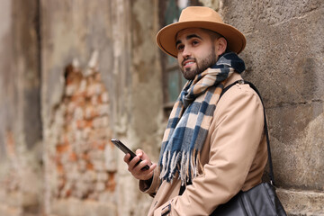 Smiling man in warm scarf with smartphone near wall outdoors. Space for text