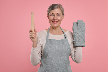 Happy housewife with oven glove and rolling pin on pink background
