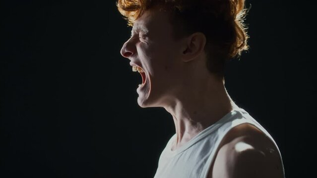 Studio shot in profile of Caucasian man in undershirt and with curly hair angrily shouting against black background