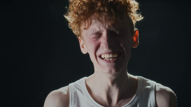 Chest up studio portrait of young man in undershirt and with ginger curly hair who cannot stop laughing against black background and looking at camera
