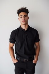 Obraz premium young Middle Eastern man standing with his hands in his pockets, wearing a black polo shirt and black pants, short curly dark hair, medium skin tone, plain white background