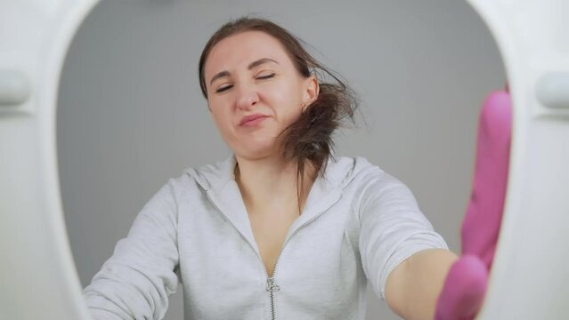 Woman wearing white hoodie is focused on cleaning toilet with brush in restroom. Using cleaning tools and products to scrub and sanitize toilet bowl and surrounding surfaces. View from toilet bottom