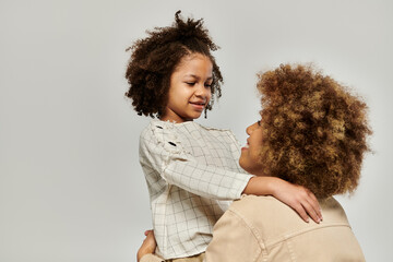 A curly African American mother and daughter wearing stylish clothes hug each other affectionately on a gray background.