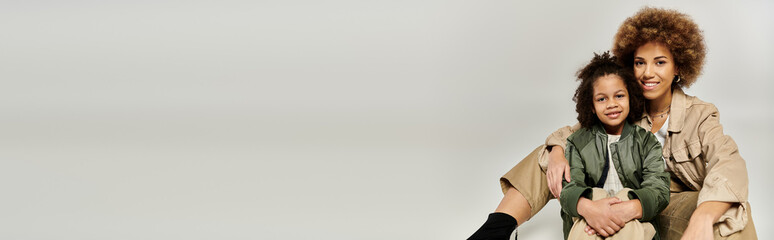 A curly African American mother and daughter in stylish clothes sit together on a white background,...