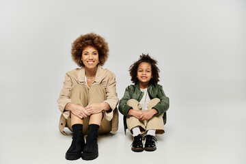 Curly African American mother and daughter sitting comfortably on a grey background in stylish...