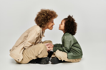 A curly African American mother and daughter in stylish clothes sit on the floor, sharing a tender...