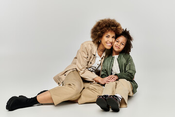 Curly African American mother and daughter in stylish attire, sitting on the floor, smiling warmly...