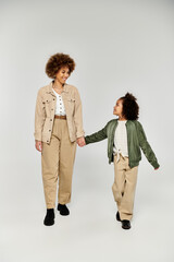 Curly African American mother and daughter holding hands in stylish outfits against a grey studio...