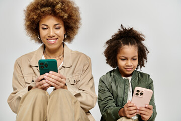 A curly African American mother and daughter in stylish clothes focus on their phones, engrossed in...