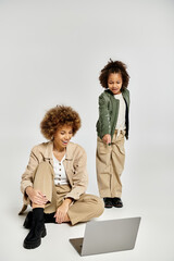 Curly African American mother and daughter sit on floor, deeply focused on laptop screen in stylish...