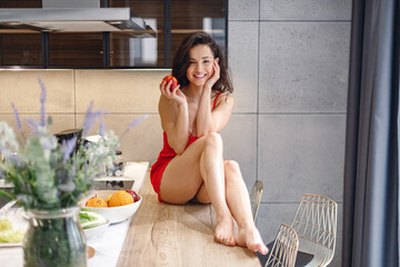 Pretty young lady sitting on the kitchen table while holding fresh apple. Woman in sexy red peignoir. Lifestyle concept