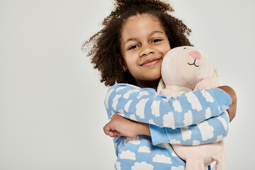 African American girl in pajamas cuddling a large stuffed animal on a grey background.