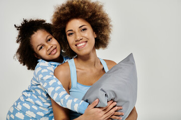 Happy African American mother and daughter in pajamas sharing a loving hug with a pillow on a grey...