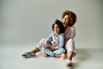 A happy African American mother and daughter sitting on the floor in cozy pajamas, sharing a...