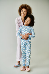 A cheerful African American mother and daughter strike a pose in matching pajamas against a grey backdrop.