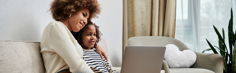 A happy African American mother and daughter sitting on a couch, engaging with a laptop together.