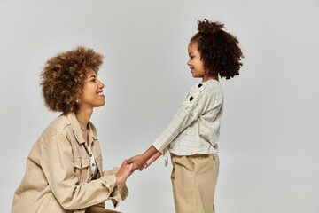 Curly African American mother and daughter holding hands in stylish clothes against a grey...