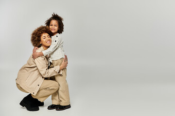 A curly African American mother warmly embraces her daughter, both in stylish attire, against a...