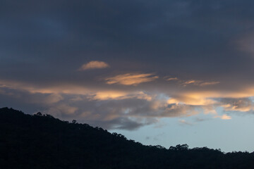 Landscape of thick clouds, orange light hitting the blue sky, silhouettes of trees, forests, and mountains. Area for text in the middle of the image