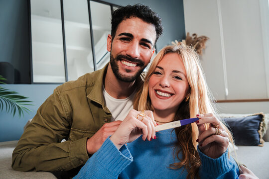 Caucasian couple holding a pregnancy test and looking at camera with a happiness expresion. Husband and wife checking the result of the maternity exam and smiling for their new baby. Parenthood