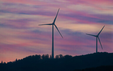 Wind turbines silhouetted against a sunset sky with purple, pink and blue hues on top of a hill