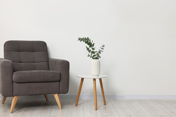 Comfortable armchair, side table and eucalyptus near white wall indoors, space for text