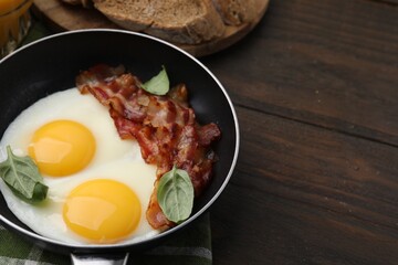 Fried eggs, bacon and basil in frying pan on wooden table, space for text