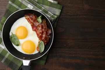 Fried eggs, bacon and basil in frying pan on wooden table, top view. Space for text