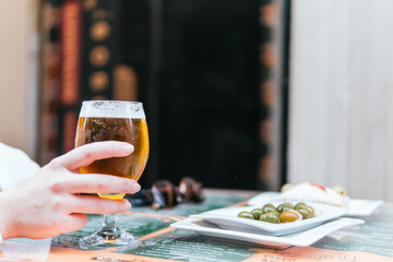 Person's hand holding a glass of beer with olives and cheese in the background