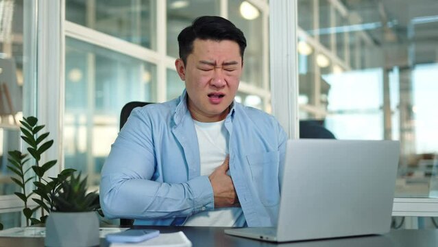 Disturbed korean entrepreneur placing hand on chest and bending with squinted face with closed eyes in personal cabinet. Sick man experiencing heart attack syndrome and feeling scared of illness.