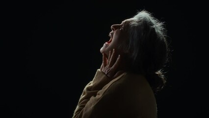 Side studio portrait in profile of senior Caucasian woman in sweater putting hands to head and yelling in pain against black background, feeling hopelessness and despair