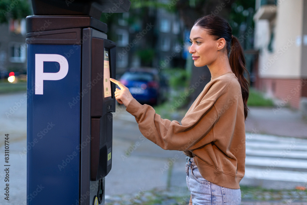 Wall mural beautiful young woman paying for outdoors parking in automated parking meter on the street, side vie