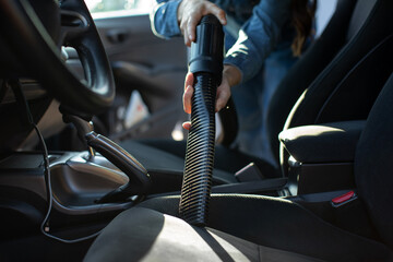 Close up of female car owner cleaning seats of vehicle with vacuum cleaner hose and attachment,...