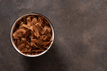 Coconut chips with cinnamon in white bowl on brown background. Tasty sugar free vegan snack. View from above. Copy space.