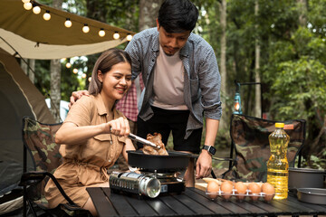 Happy Asian couple cooking food in a pan on tourist gas burner near the tent at camp. Camping and...