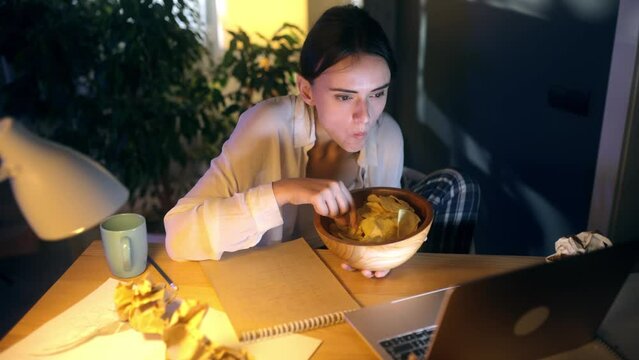 Tired young woman after hard work day eating chips but watching boring uninteresting movie show series on laptop at home workplace Exhausted female feeling sad about senseless program indoors