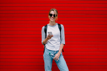 Full length portrait of cheerful female standing with smartphone and water bottle on urban red...
