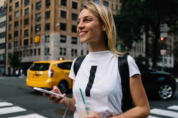 Cheerful young female tourist walking on city street passing cars and crosswalk enjoying big city,smiling hipster girl excited with fast taxi service calling cab via application on smartphone