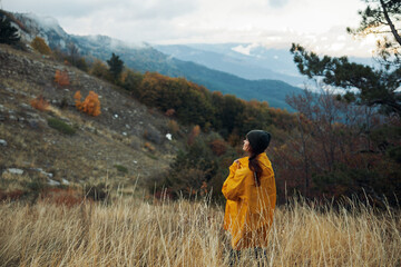 Woman in a vibrant yellow raincoat standing in a scenic field with majestic mountains in the background Travel adventure in nature beauty