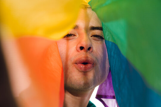 Close-up front view of Latin Gay Man looking at camera blowing a kiss covered by a pride flag