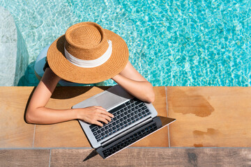 Young Asian traveler woman working on computer at pool in luxury resort during her summer holiday. Work from anywhere, Technology and lifestyle concept, copy space