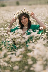Fototapeta premium Happy woman in a field of daisies with a wreath of wildflowers on her head. woman in a green dress in a field of white flowers. Charming woman with a bouquet of daisies, tender summer photo
