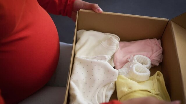 Unrecognizable pregnant African American woman girl female mother pregnancy at home couch unpack box parcel unpacking order hold baby shoes socks put on belly delivery cloth shopping clothes prepare
